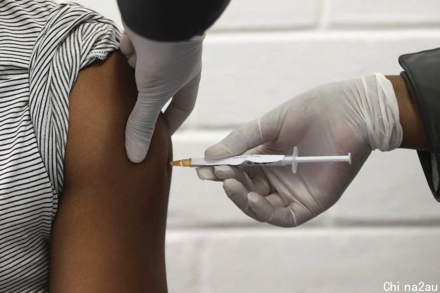 The shoulder of a volunteer is seen as they receive an injection by a person wearing gloves.