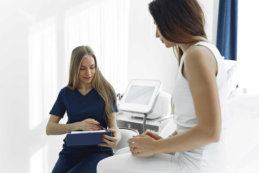 A woman wearing navy scrubs writes on a clipboard while another woman sits on a hospital bed.