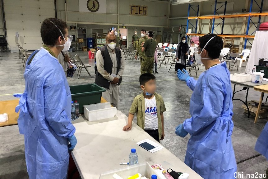 People wearing masks walk toward medical professionals wearing blue gowns and masks in a large hangar space