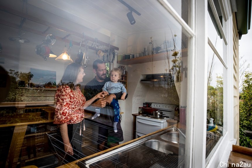 A mother and father play with their baby boy. The photo has been taken from outside through their kitchen window.