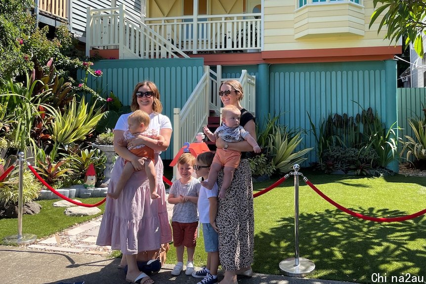 Two smiling women with young children stand outside a small yellow and red cottage