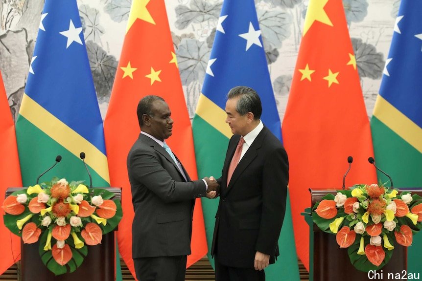 In front of a row of Solomon Islands and Chinese flags, Jeremiah Manele and Wang Yi shake hands in between lecterns.