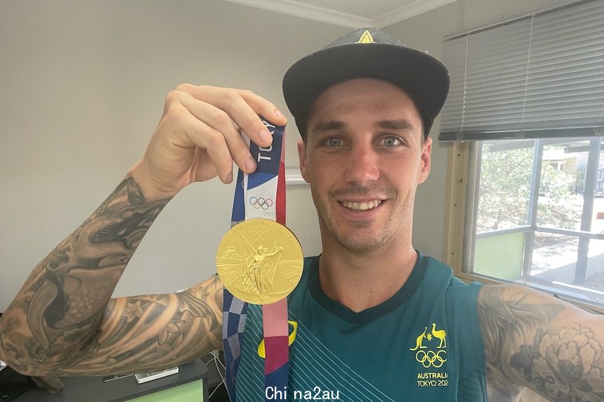 A man with a lot of tattoos in a singlet top and cap holds a gold medal and smiles as he takes a selfie