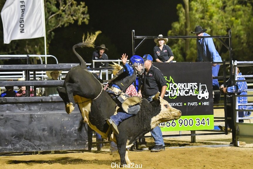 Bonnie on top of a black bucking bull wearing protective clothing and a helmet with one hand in the air