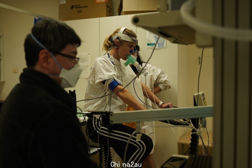 A woman on an exercise bike wearing breathing apparatus, with healthcare professionals on either side of her. 