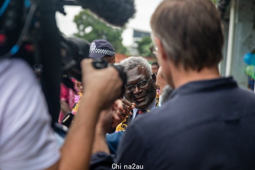 The Prime Minister holds up a finger. He's surrounded by people, including in the foreground and journalist and camera operator.