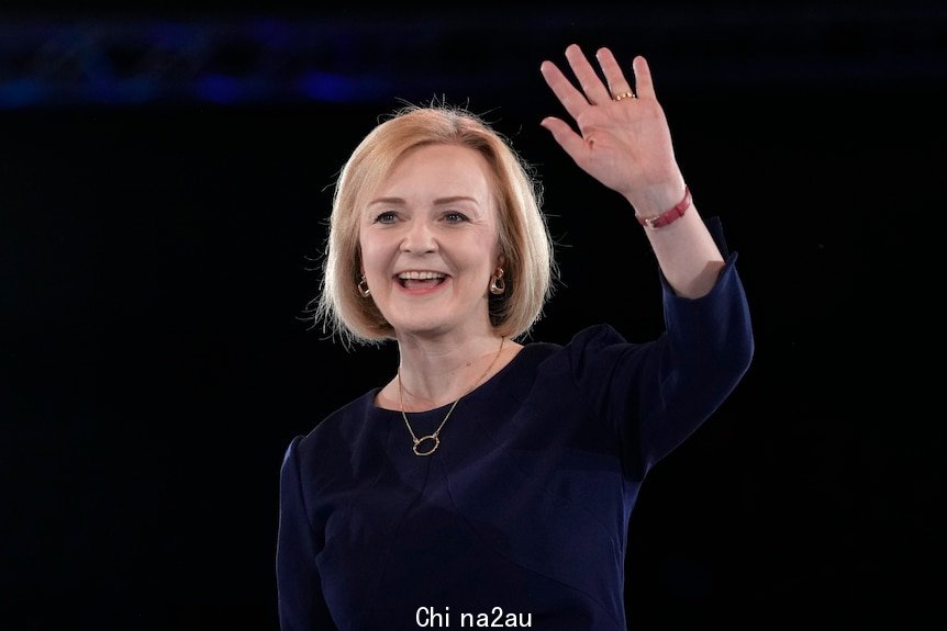 Liz Truss, in a navy dress, smiles and waves against a dark background. She wears a red strapped wristwatch