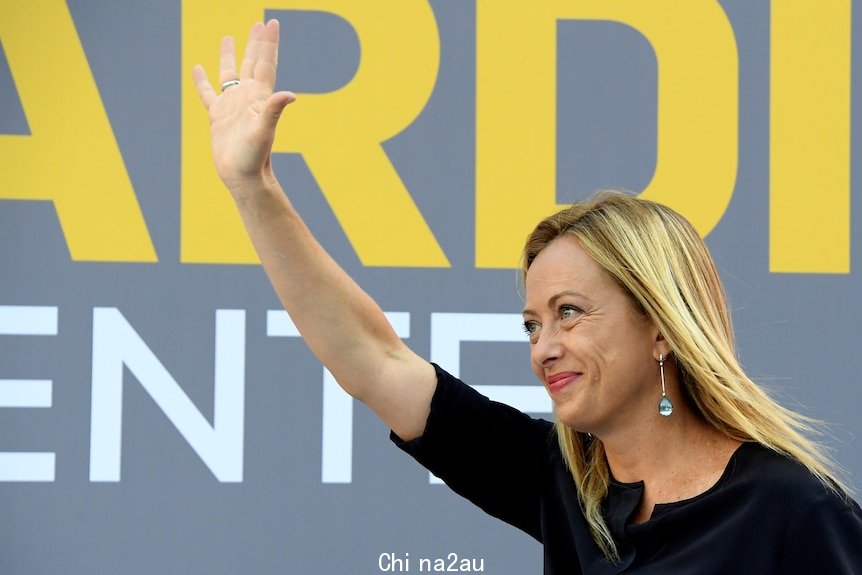 A woman with blonde hair wearing a black dress waves in front of a grey background with yellow and white writing. 