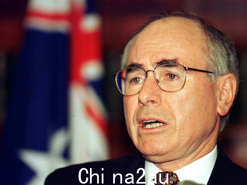 John Howard，浓密的黑眉毛和他标志性的眼镜，站在澳大利亚国旗前。