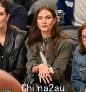 在纽约市的一场篮球比赛中，卡莉·克劳斯 (Karlie Kloss) 坐在场边座位上与朋友击掌，身着卡其色夹克，展现出时尚的身材